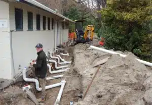 Worker installing underground drainage pipes beside a building, with multiple PVC pipes connected along the wall, trench excavation in progress, and a mini excavator in the background.